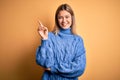 Young beautiful blonde woman wearing turtleneck sweater over yellow isolated background with a big smile on face, pointing with Royalty Free Stock Photo