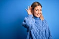 Young beautiful blonde woman wearing winter wool sweater over blue isolated background smiling with hand over ear listening an Royalty Free Stock Photo