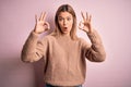 Young beautiful blonde woman wearing winter wool sweater over pink isolated background looking surprised and shocked doing ok Royalty Free Stock Photo