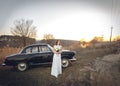 Young beautiful bride is next near to black retro car sunset background. Wedding with vintage old retro car. woman holds beautiful Royalty Free Stock Photo