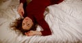 A young beautiful brown-haired girl in a red dress lies on a crumpled white bed, moves, turns over, poses, crosses her Royalty Free Stock Photo