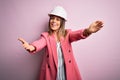 Young beautiful brunette architect woman wearing safety helmet over pink background looking at the camera smiling with open arms Royalty Free Stock Photo