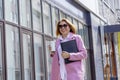 a young beautiful brunette businesswoman in sunglasses and a pink coat is walking along a city street Royalty Free Stock Photo