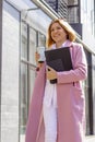 a young beautiful brunette businesswoman in a white blouse and light trousers and a pink coat is walking along a city street, in Royalty Free Stock Photo