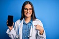 Young beautiful brunette doctor woman wearing glasses and coat holding smartphone with surprise face pointing finger to himself Royalty Free Stock Photo