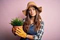 Young beautiful brunette farmer woman wearing apron and hat holding pot with plants with a confident expression on smart face Royalty Free Stock Photo