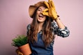 Young beautiful brunette farmer woman wearing apron and hat holding pot with plants with happy face smiling doing ok sign with Royalty Free Stock Photo