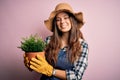 Young beautiful brunette farmer woman wearing apron and hat holding pot with plants with a happy face standing and smiling with a Royalty Free Stock Photo