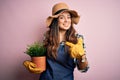 Young beautiful brunette farmer woman wearing apron and hat holding pot with plants with surprise face pointing finger to himself Royalty Free Stock Photo