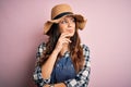 Young beautiful brunette farmer woman wearing apron and hat over pink background with hand on chin thinking about question,