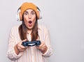 Young beautiful brunette gamer woman playing video game using joystick and headphones scared and amazed with open mouth for