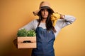 Young beautiful brunette gardener woman wearing apron and hat holding box with plants with angry face, negative sign showing Royalty Free Stock Photo