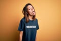 Young beautiful brunette girl doing volunteering wearing t-shirt with volunteer message word sticking tongue out happy with funny