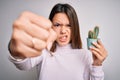 Young beautiful brunette girl holding small cactus plant pot over isolated white background annoyed and frustrated shouting with Royalty Free Stock Photo