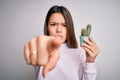 Young beautiful brunette girl holding small cactus plant pot over isolated white background pointing with finger to the camera and Royalty Free Stock Photo