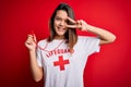 Young beautiful brunette lifeguard girl wearing t-shirt with red cross using whistle Doing peace symbol with fingers over face,