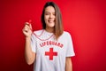 Young beautiful brunette lifeguard girl wearing t-shirt with red cross using whistle with a happy and cool smile on face