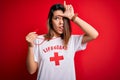 Young beautiful brunette lifeguard girl wearing t-shirt with red cross using whistle making fun of people with fingers on forehead