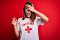 Young beautiful brunette lifeguard girl wearing t-shirt with red cross using whistle peeking in shock covering face and eyes with
