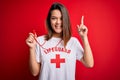 Young beautiful brunette lifeguard girl wearing t-shirt with red cross using whistle pointing finger up with successful idea