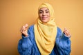 Young beautiful brunette muslim woman wearing arab hijab over isolated yellow background doing money gesture with hands, asking Royalty Free Stock Photo