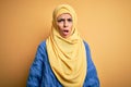 Young beautiful brunette muslim woman wearing arab hijab over isolated yellow background In shock face, looking skeptical and