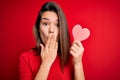 Young beautiful brunette romantic girl holding red paper heart shape over isolated background cover mouth with hand shocked with Royalty Free Stock Photo