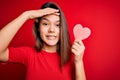 Young beautiful brunette romantic girl holding red paper heart shape over isolated background stressed with hand on head, shocked Royalty Free Stock Photo