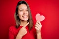 Young beautiful brunette romantic girl holding red paper heart shape over isolated background very happy pointing with hand and Royalty Free Stock Photo