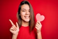 Young beautiful brunette romantic girl holding red paper heart shape over isolated background very happy pointing with hand and Royalty Free Stock Photo