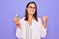 Young beautiful brunette scientist woman wearing coat and glasses holding test tube pointing thumb up to the side smiling happy Royalty Free Stock Photo