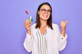 Young beautiful brunette scientist woman wearing coat and glasses holding test tube pointing thumb up to the side smiling happy Royalty Free Stock Photo