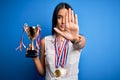 Young beautiful brunette successful woman wearing medals holding trophy with open hand doing stop sign with serious and confident