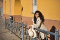 Young, beautiful, brunette woman with curly hair and beige coat and hat, leaning on a railing looking at camera smiling and happy Royalty Free Stock Photo