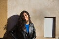 Young, beautiful, brunette woman with curly hair and black leather jacket and pants, leaning against a wall receiving the sun's Royalty Free Stock Photo