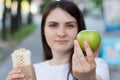 Young beautiful brunette woman on a diet chooses food - what to eat an apple or fast food shawarma. Bites the apple and