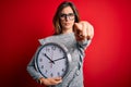 Young beautiful brunette woman doing countdown holding big clock over red background pointing with finger to the camera and to Royalty Free Stock Photo