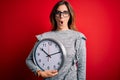 Young beautiful brunette woman doing countdown holding big clock over red background scared in shock with a surprise face, afraid Royalty Free Stock Photo