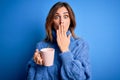 Young beautiful brunette woman drinking cup of coffe over isolated blue background cover mouth with hand shocked with shame for Royalty Free Stock Photo