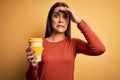 Young beautiful brunette woman drinking cup of takeaway coffe over yellow background stressed with hand on head, shocked with Royalty Free Stock Photo