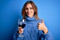 Young beautiful brunette woman drinking glass of red wine over  blue background happy with big smile doing ok sign, thumb Royalty Free Stock Photo