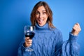 Young beautiful brunette woman drinking glass of red wine over isolated blue background screaming proud and celebrating victory Royalty Free Stock Photo