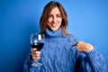 Young beautiful brunette woman drinking glass of red wine over isolated blue background with surprise face pointing finger to Royalty Free Stock Photo