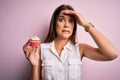 Young beautiful brunette woman eating chocolate cupcake over isolated pink background stressed with hand on head, shocked with Royalty Free Stock Photo