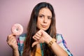 Young beautiful brunette woman eating sweet pink doughnut over isolated background with angry face, negative sign showing dislike