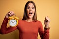 Young beautiful brunette woman holding alarm clock over isolated yellow background screaming proud and celebrating victory and Royalty Free Stock Photo