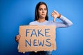 Young beautiful brunette woman holding banner with fake news message with angry face, negative sign showing dislike with thumbs