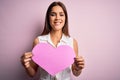 Young beautiful brunette woman holding big pink heart paper celebrating valentine day with a happy face standing and smiling with Royalty Free Stock Photo