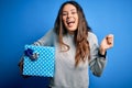 Young beautiful brunette woman holding birthday gift over isolated blue background screaming proud and celebrating victory and Royalty Free Stock Photo