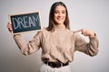 Young beautiful brunette woman holding blackboard with dream message word with surprise face pointing finger to himself